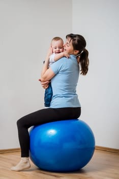 Mother exercises with blue fit ball at home holding her baby on hands