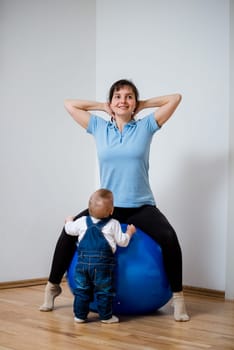 Mother exercises with big blue fit ball at home and her baby is watching her