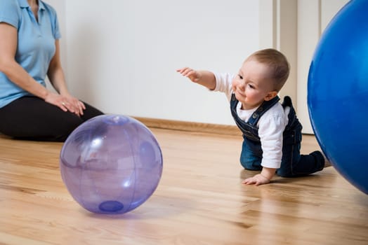 Baby plays with inflatable ball at home on floor, mother in background