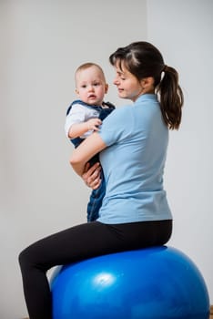 Mother exercises with blue fit ball at home holding her baby on hands