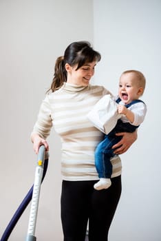 Cleaning up the room - mother with vacuum cleaner holding her baby on hands