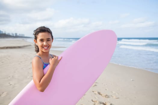 Cute smiling young girl holding pink surfboard at the beach