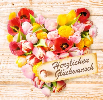 German birthday wishes on a colourful floral heart formed of fresh spring tulips in vibrant colours arranged on a rustic wooden background with a tag saying - Happy Birthday or Herzlichen Gluckwunsch