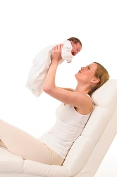 An adult woman with a white shirt lies on a white couch and holds an infant dressed in white in front of her in the air,exposed against a white background.