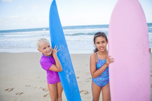 Two cute smiling young girls holding surfboards upright at the beach with ocean in background