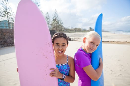 Two cute smiling young girls holding surfboards upright at the beach
