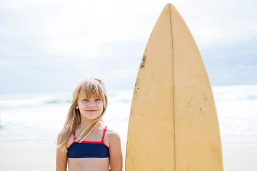 Happy smiling cute young girl standing next to surfboard on beach with sky and ocean in background