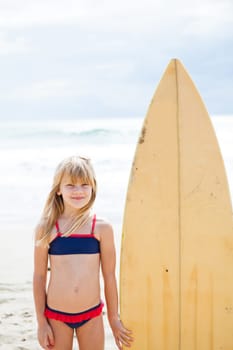 Happy smiling cute young girl standing next to surfboard on beach with sky and ocean in background