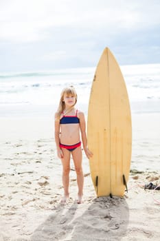 Cute young girl standing next to surfboard on beach with ocean in background