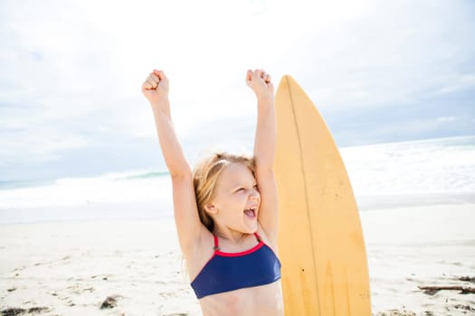 Excited happy young girl with arms raised on beach with surfboard and ocean and sky in background