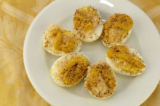 Three prepared boiled eggs halved with yolk prepared with deviled egg mixture and placed on a round white plate with a yellow gold floral place mat background..