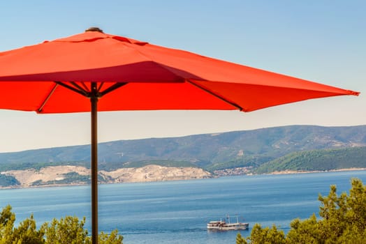 red terrace,garden umbrella against blue sea and cloudless sky,visible coastline,ship in distance.