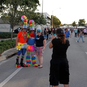 WILTON MANORS, FLORIDA - JUNE 22, 2013: Two adult people in colorful costumes with big hats pose next to a gay pride celebration attendee while another person makes a photograph on a smart phone.