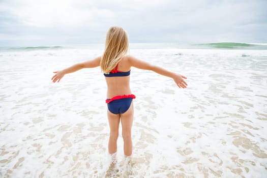 Cute young girl standing in water at beach facing the ocean with arms raised