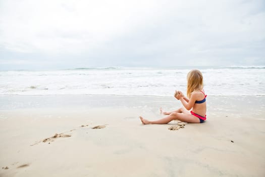 Young blond girl sitting on sand and playing at beach with ocean in background