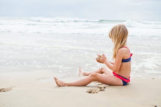 Cute young girl sitting on sand and playing at beach with ocean in background