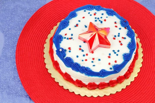 Round Independence Day holiday cake decorated with white frosting, red and blue ribbons, edible stars and a red plastic star on the center top. The cake is on a gold base, then red and blue placemats.