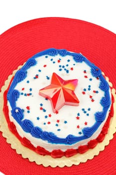 Festive red, white and blue July 4th cake, decorated with edible star sprinkles, one large plastic star, and frosting. The dessert is placed on a gold foundation over a round red place mat..