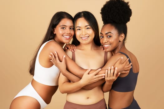 Three young women of different backgrounds smiling at camera, confidently pose and embrace in underwear, showcasing natural beauty on beige background.