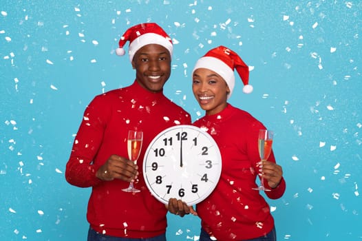 Happy African American Couple In Santa Hats Holding Champagne Glasses And Large Clock Amidst Falling Confetti On Blue Studio Background, Waiting Midnight Celebration And New Year's Eve Festivities