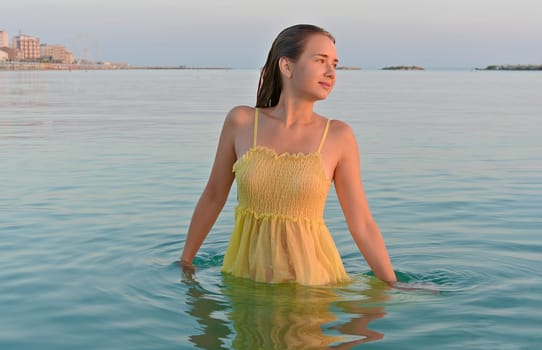 Young woman in a yellow dress under sunrise on the seashore against the backdrop of the city.