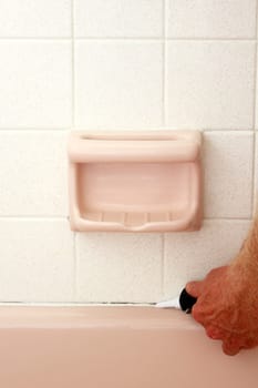 Caucasian hand of an adult man applying white mold resistant caulk to repair above the rim of a pink bath tub over an opened seam below speckled white tile wall of an old home to prevent water damage..