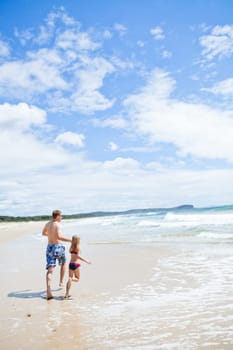 Father and young daughter holding hands and running along beach at edge of water
