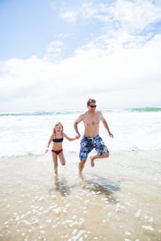 Happy smiling father and young daughter holding hands and running towards beach away from waves