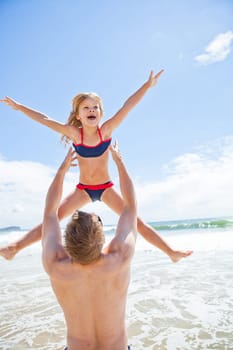 Father throwing happy smiling young daughter in air at beach with water and sky in background