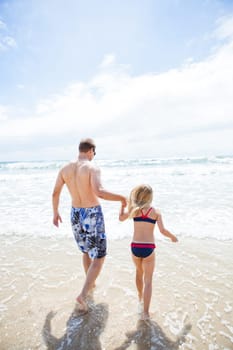 Father and young daughter holding hands and walking into water together at beach