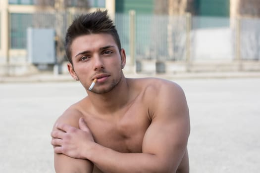 Shirtless muscular young man smoking cigarette outside looking at camera