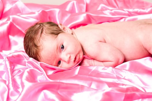 Newborn baby (6 weeks) lies on a pink satin blanket and looks cute into the camera.
