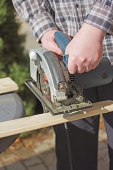 Closeup view of a man in a checkered shirt that is cutting wooden board by electric jigsaw outdoors.