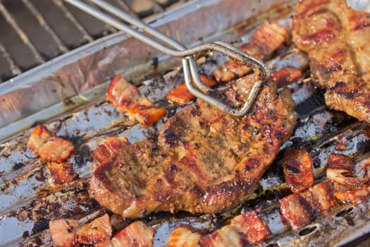 Grilled steak and bacon on an aluminum tray, which prevents dripping fat into the fire.