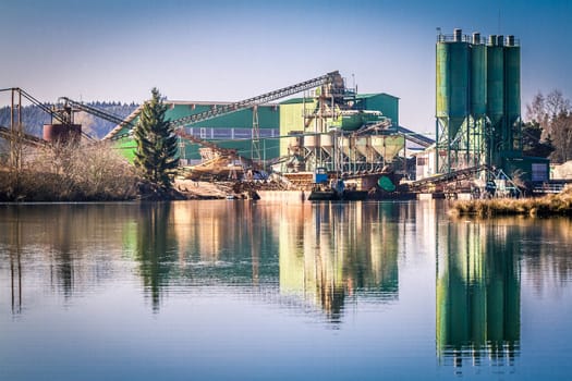 A green-painted concrete plant on a sunny spring day is reflected in a dredging lake.