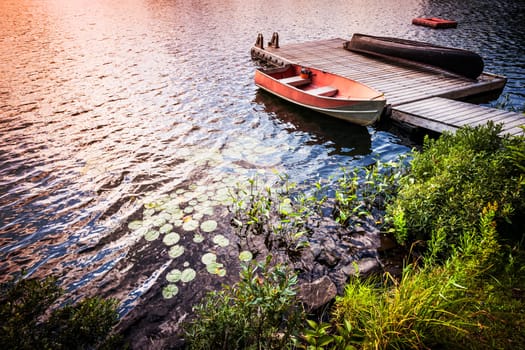 Sunset reflecting in lake water with rowboat tied to wooden dock at rocky shore. Ontario, Canada.