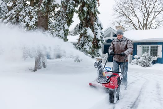 Man using snowblower to clear deep snow on driveway near residential house after heavy snowfall.