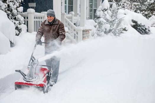Man using snowblower to clear deep snow on driveway near residential house after heavy snowfall.