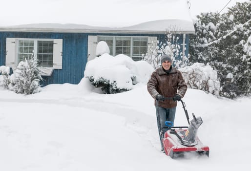 Man using snowblower to clear deep snow on driveway near residential house after heavy snowfall.