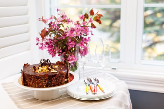 Round gourmet chocolate cake on table next to window with plates, cutlery, and pink spring bouquet