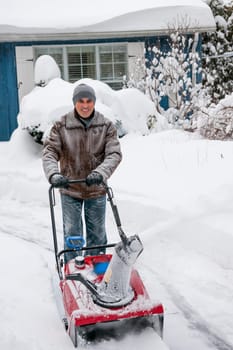 Man using snowblower to clear deep snow on driveway near residential house after heavy snowfall.