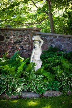 Lush green summer garden with perennial plants and statue near stone wall