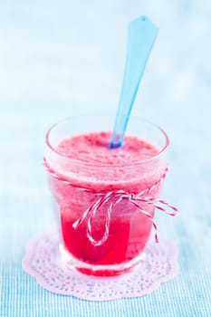Closeup of strawberry smoothie and blue plastic spoon in glass on pink mat resting on light blue tablecloth