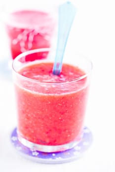 Closeup of strawberry smoothie and blue plastic spoon in glass on purple plate with another in background