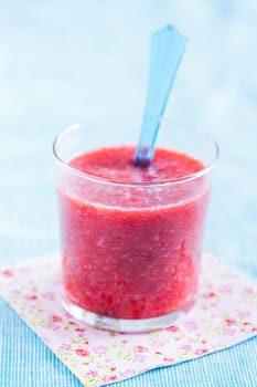 Closeup of strawberry smoothie and blue plastic spoon in glass resting on floral serviette on light blue tablecloth
