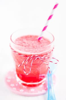 Closeup of strawberry smoothie and striped straw in glass on pink plate with blue plastic spoon