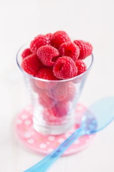Closeup of fresh red raspberries in glass with blue plastic spoon resting on small pink plate on light surface