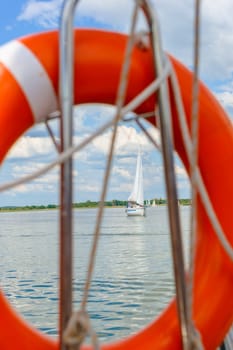 Sailing boat in lake, photo taken through orange lifebelt, rigging, white clouds in blue sky.