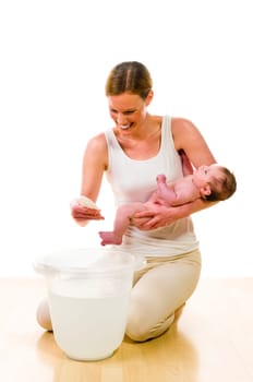 Adult mother wearing bright casual clothes, she kneels with her newborn baby on the floor, in front of a bathing bucket. In her hand she holds a thermometer and checked the termperature of the bath water. Isolated against white background.