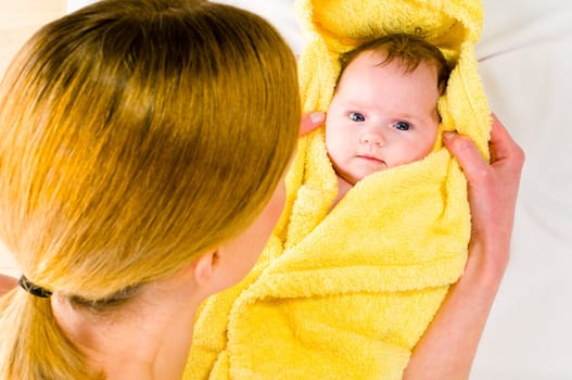 Adult mother wearing bright casual clothes, she kneels on the floor and dries the freshly bathed newborn baby with a yellow towel and smiling while doing. Isolated against white background.